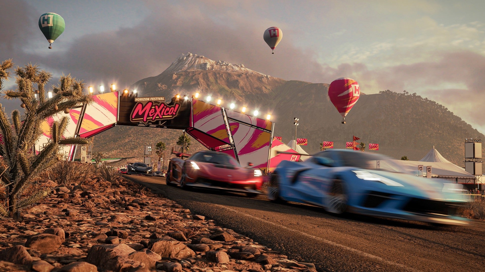 Two race cars on a desert track with hot air balloons and mountains in the background.
