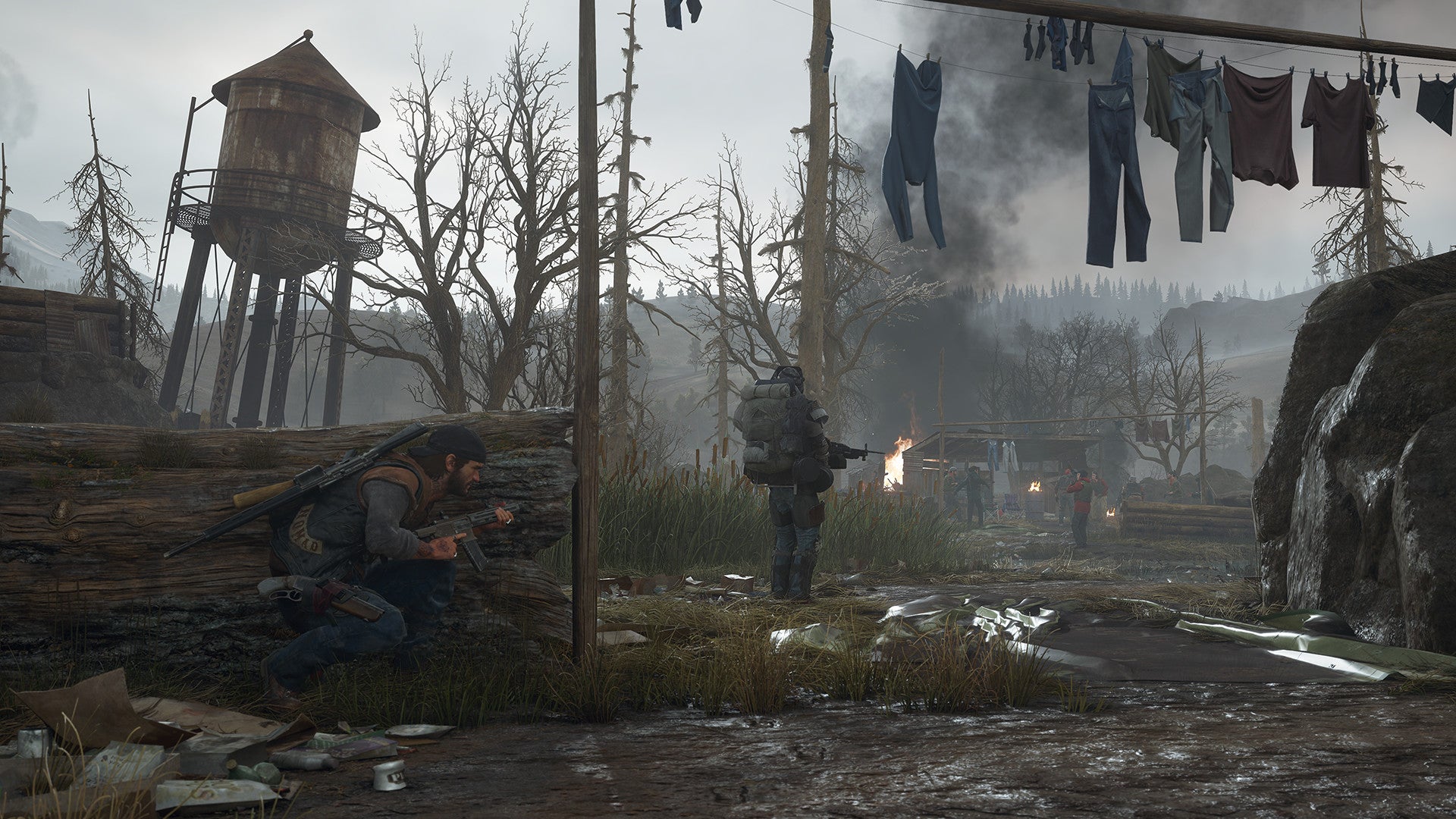 Two characters in a post-apocalyptic setting with a wooden structure, clothes hanging, and a fire in the background.