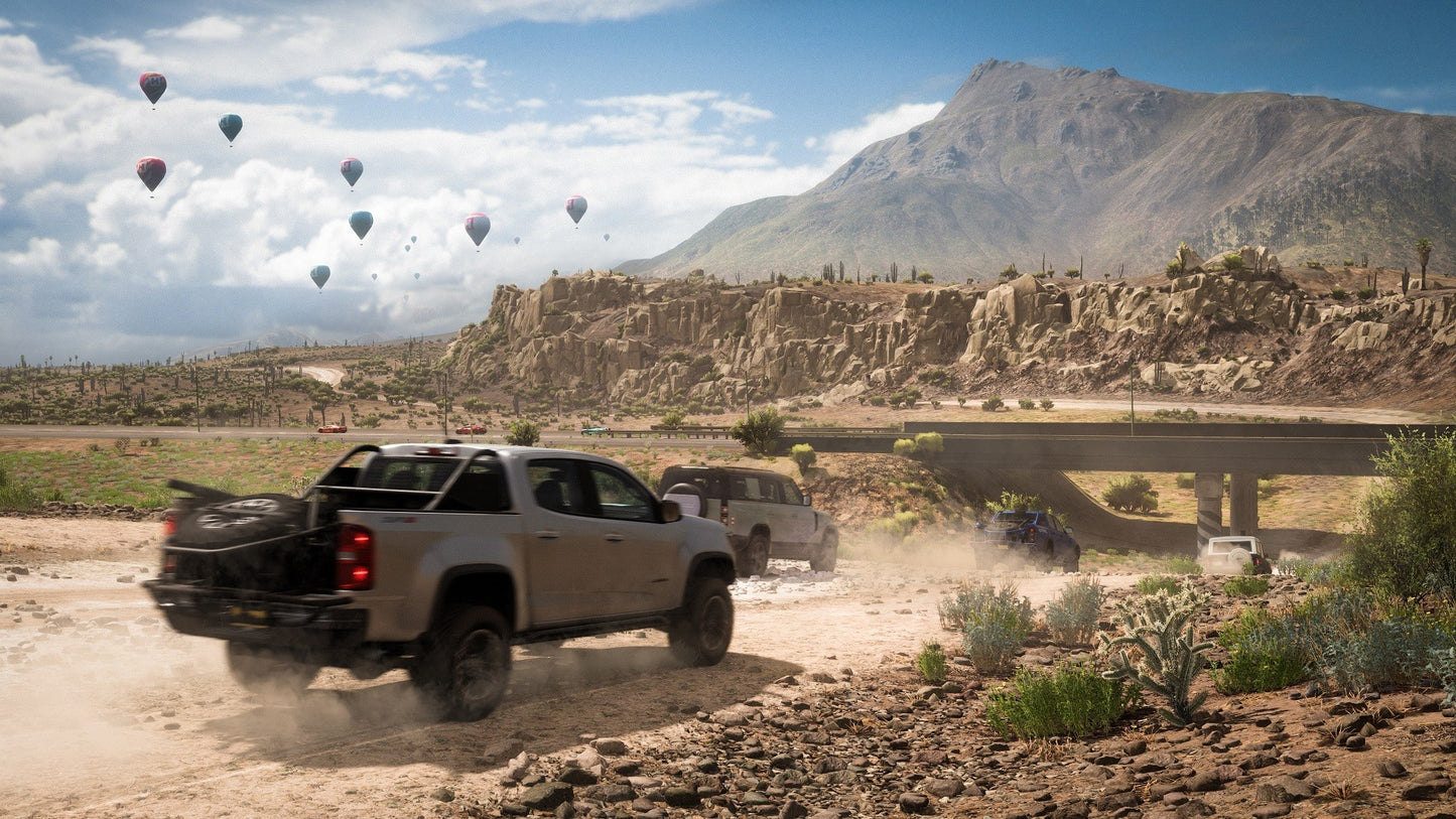 Two trucks driving on a dirt road with hot air balloons and mountains in the background.