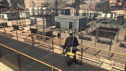 Person walking on a platform in a desert-like industrial area with buildings and equipment.